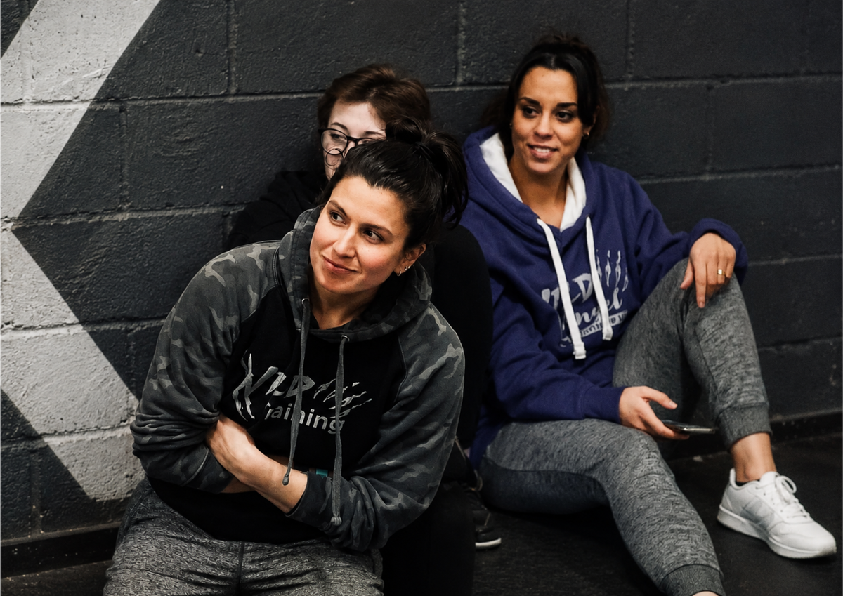 Three women wearing Pathfinder-branded hoodies sit against a gym wall while watching an MMA event. The candid moment captures friendship, support and the community spirit that exists beyond the competition itself.
