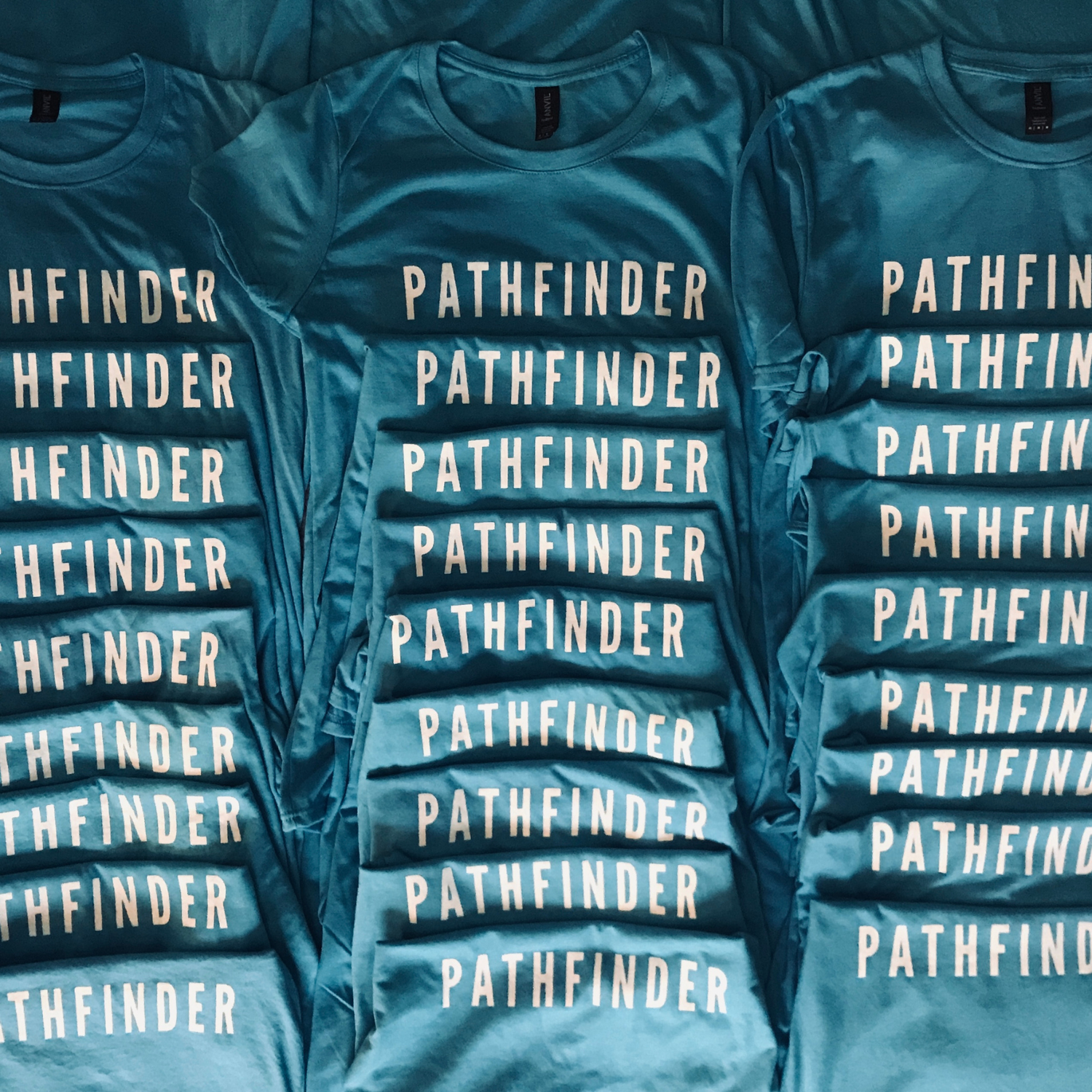 Stack of blue t-shirts with 'PATHFINDER' printed on them. Pathfinder Printworks hand screen print clothing for club, teams and businesses.