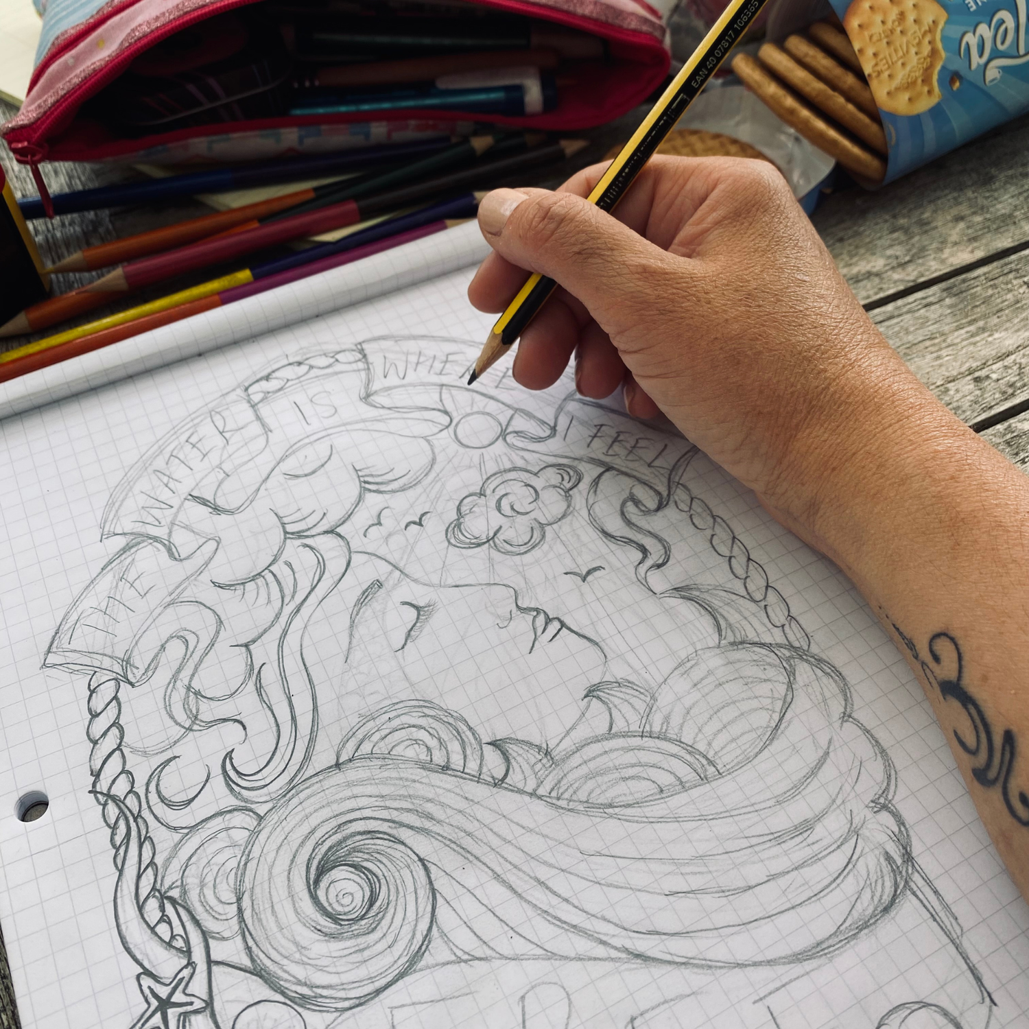 Person drawing a detailed illustration of a mermaid with a pencil on a notebook. Pathfinder Printworks hand screen print clothing for club, teams and businesses.