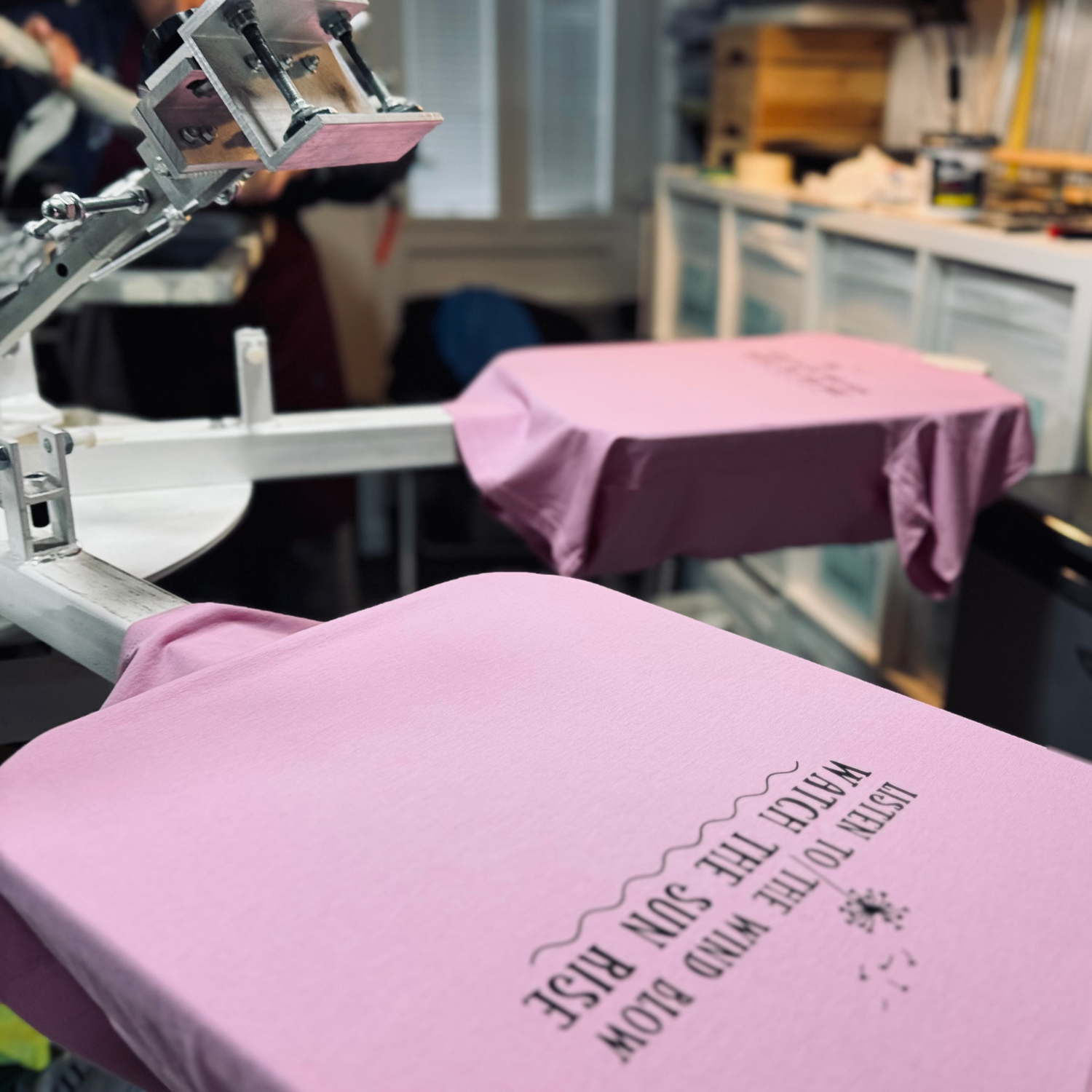 Pink t-shirt with "Listen to the wind blow" text being printed on a screen printing machine. Pathfinder Printworks hand screen print for clubs, teams and businesses.
