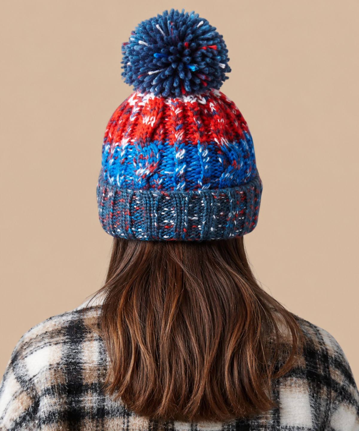 A person shown from the back wearing a chunky knit pom-pom beanie with horizontal stripes of navy blue, red, and white, paired with a plaid shirt against a solid beige background. Pathfinder Printworks offer embroidery services to teams, clubs and businesses.