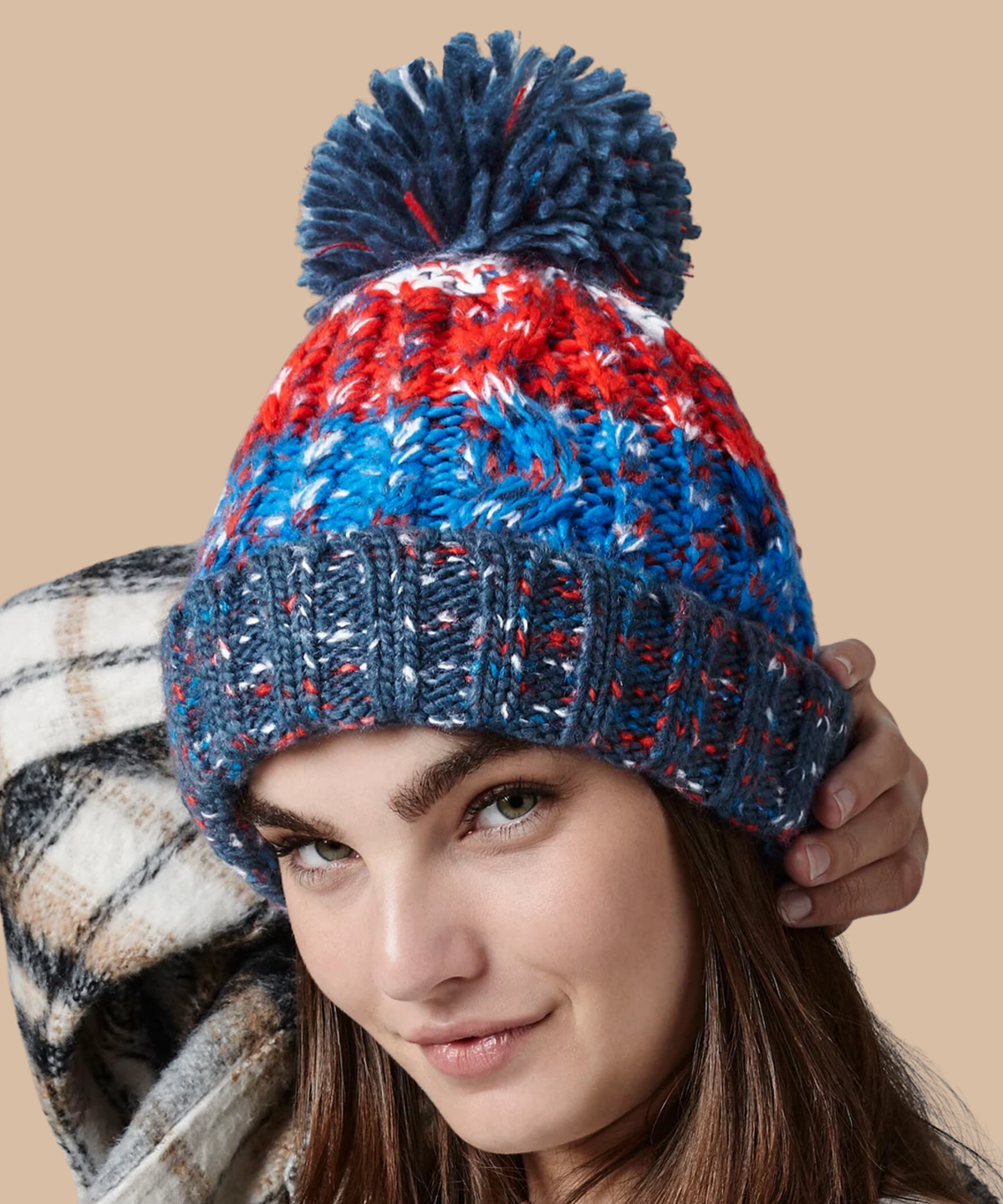 A woman wearing a chunky knit pom-pom beanie with horizontal stripes of navy blue, bright blue, red, and white, against a solid beige background. Pathfinder Printworks offer embroidery services to teams, clubs and businesses.