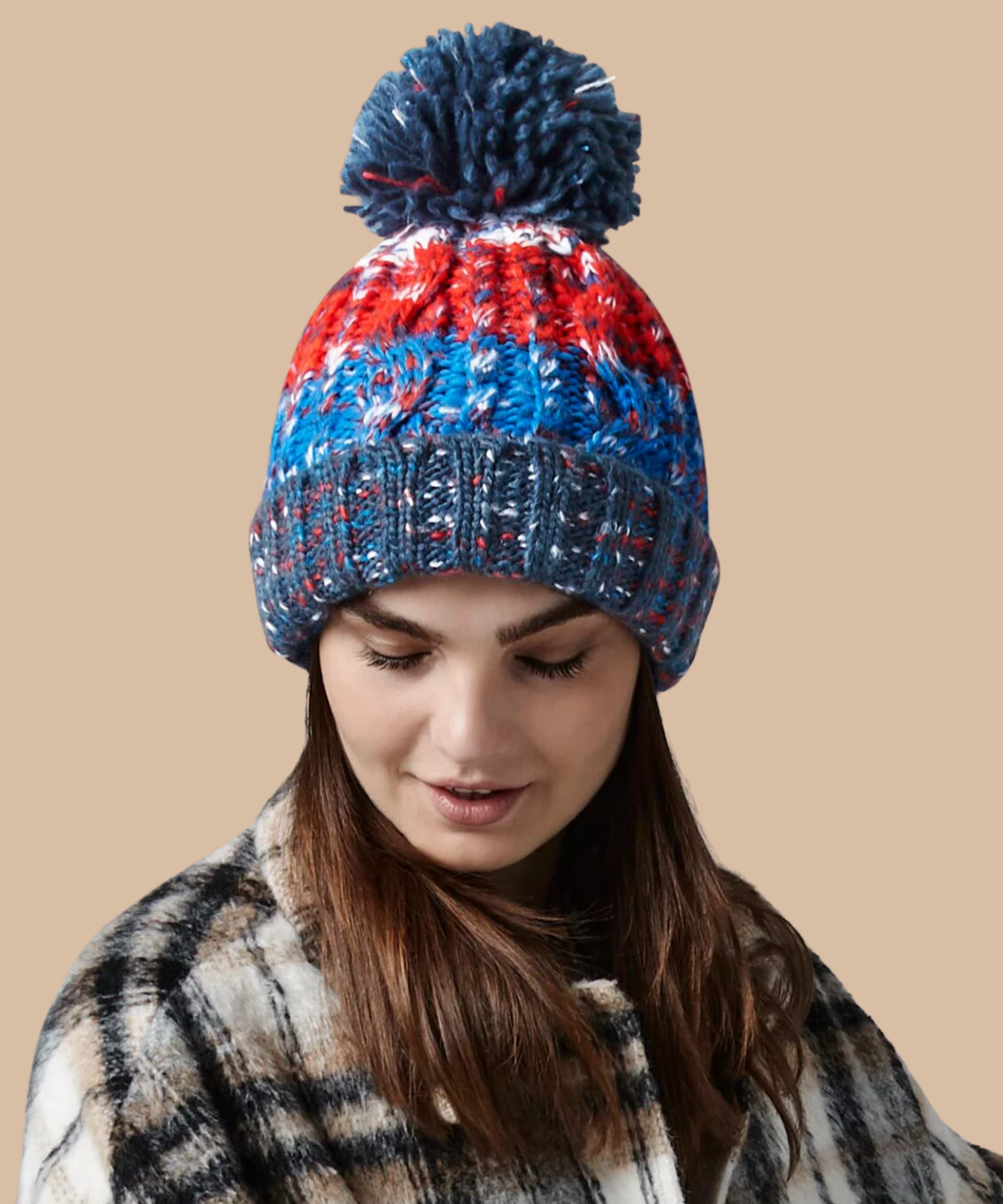 A woman wearing a corkscrew knitted pom-pom beanie with stripes of navy, red, and blue, paired with a plaid coat against a solid beige background. Pathfinder Printworks offer embroidery services to teams, clubs and businesses.