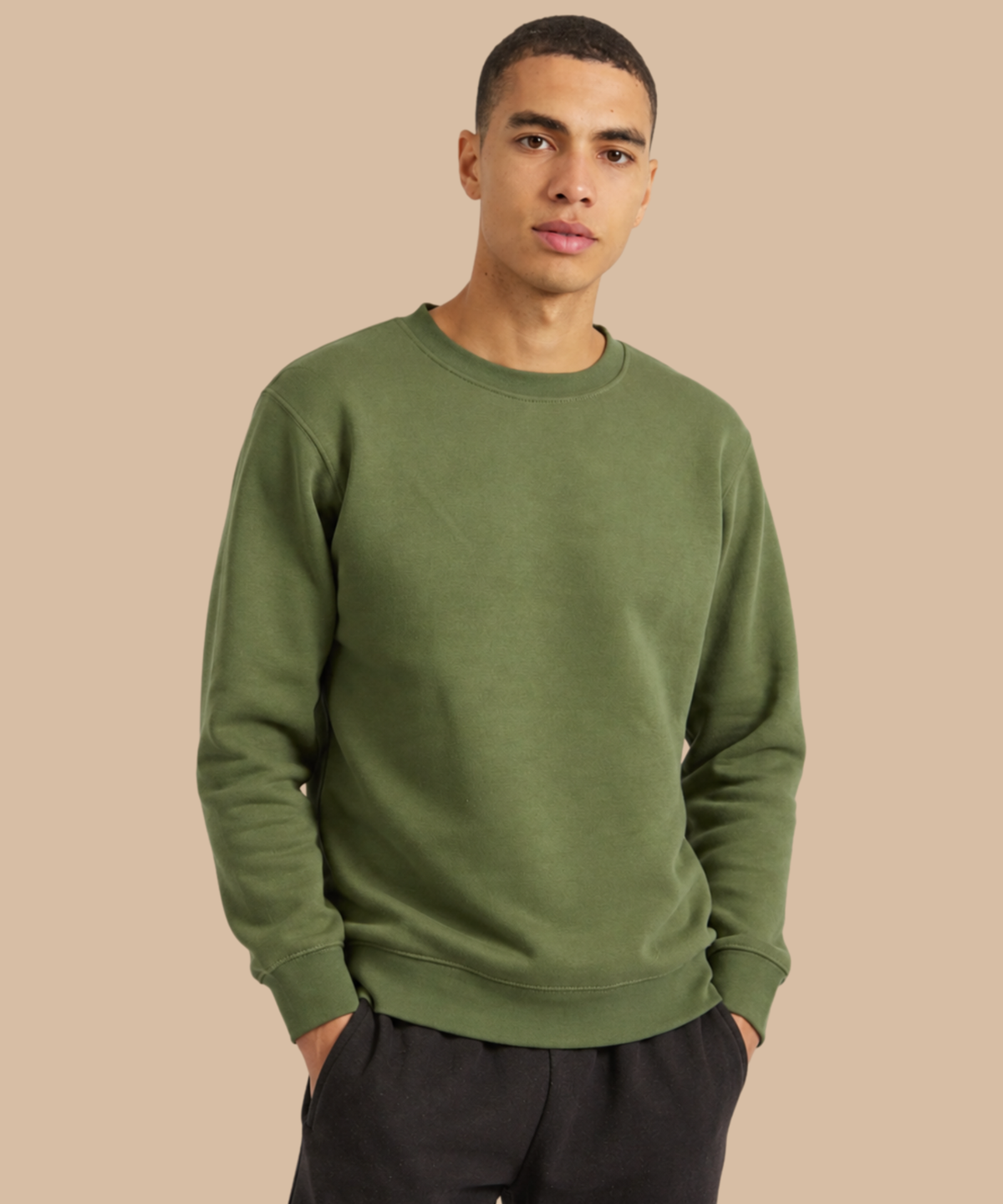 A man wearing an olive green crewneck sweatshirt and black pants, standing against a solid beige background. Pathfinder Printworks provides printing and embroidery services to teams, clubs and businesses - based in South Wales.