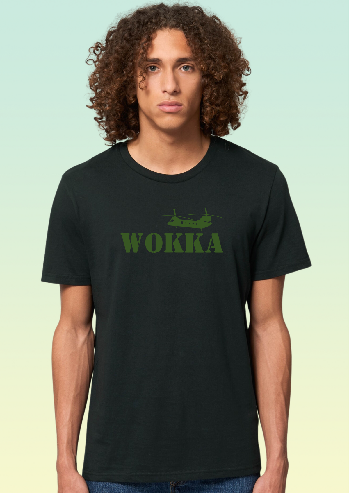 A man with long curly brown hair wearing a black crewneck t-shirt with a green graphic of a helicopter and the word 'WOKKA' printed on the chest, set against a light green gradient background. Exclusive to Flying Teez.