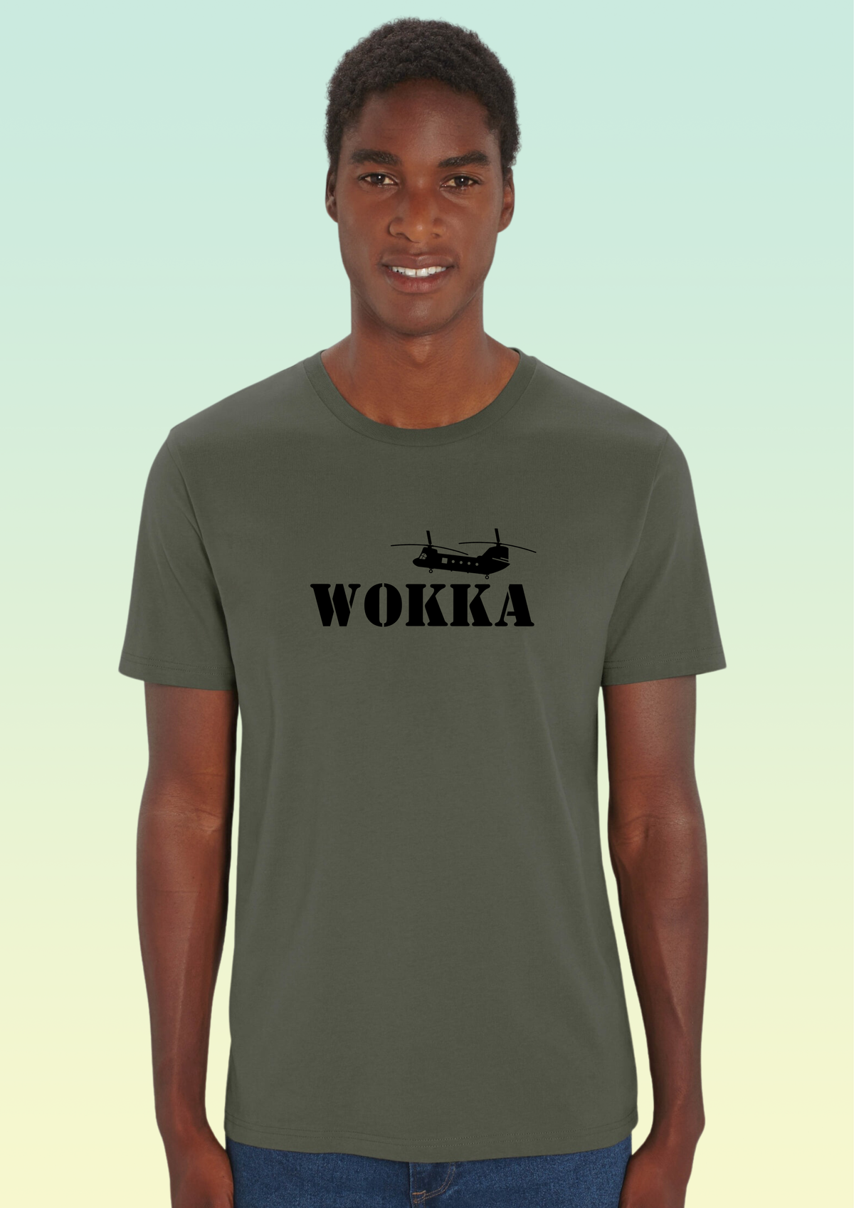 A man wearing an olive green crewneck t-shirt with a black graphic of a helicopter and the word 'WOKKA' printed on the chest, set against a light green gradient background. Exclusive to Flying Teez.
