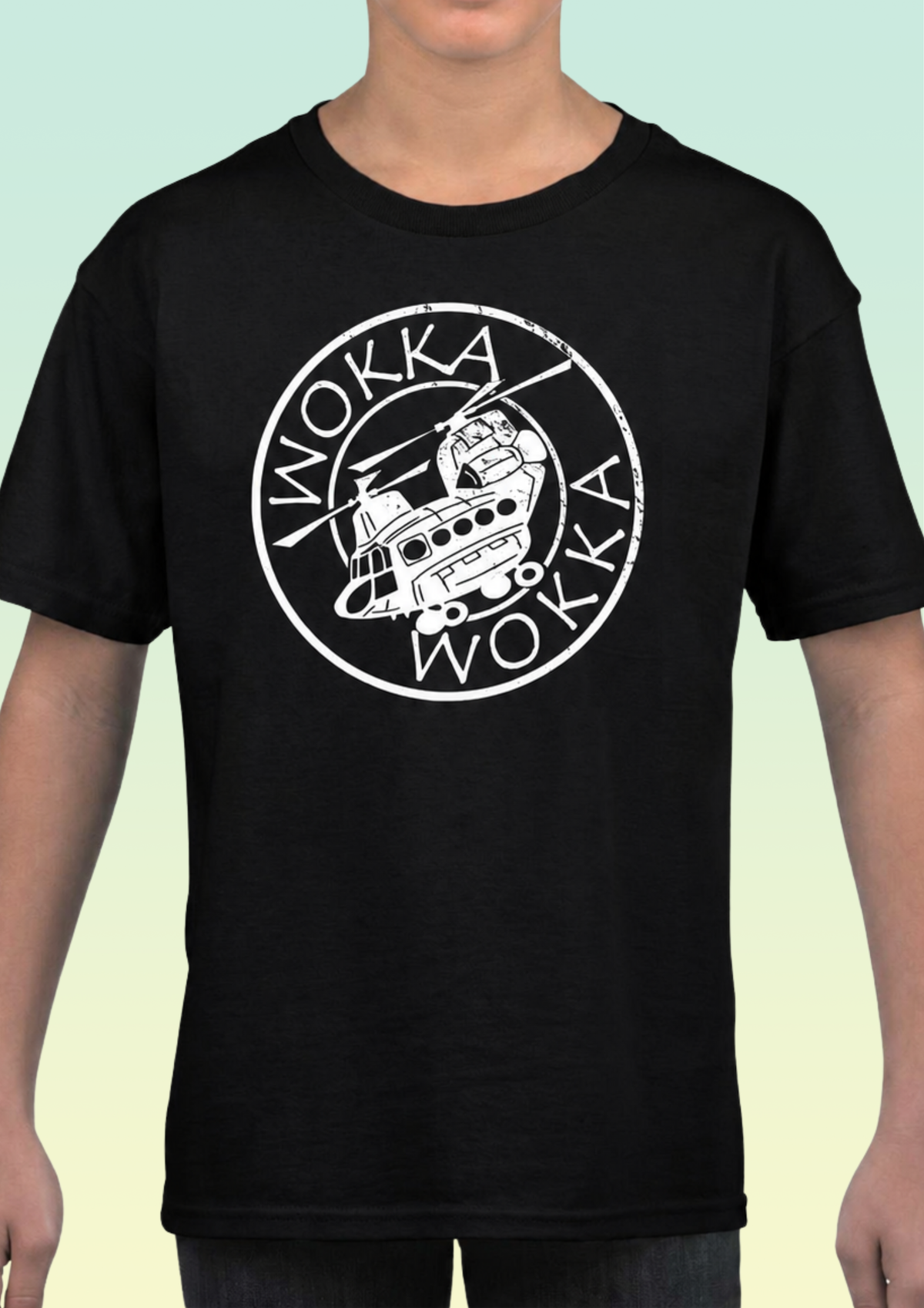 A person wearing a black short-sleeve T-shirt with a large white circular graphic on the chest featuring the word 'WOKKA' and a helicopter illustration, against a pale green-to-yellow gradient background. Exclusive to Flying Teez.