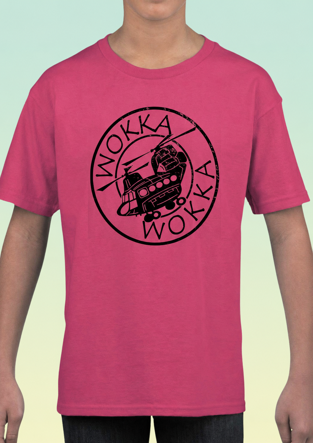 A person wearing a bright pink short-sleeve t-shirt with a black circular 'Wokka Wokka' helicopter graphic printed on the chest, set against a light mint green gradient background. Exclusive to Flying Teez.