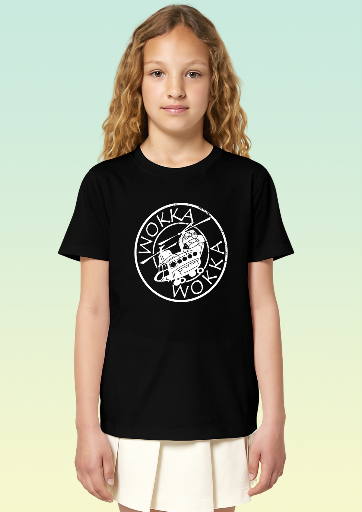 A young girl with long curly blonde hair stands against a light green-to-yellow gradient background, wearing a black short-sleeve t-shirt with a white circular Chinook helicopter graphic on the chest and a cream pleated skirt. Exclusive to Flying Teez.