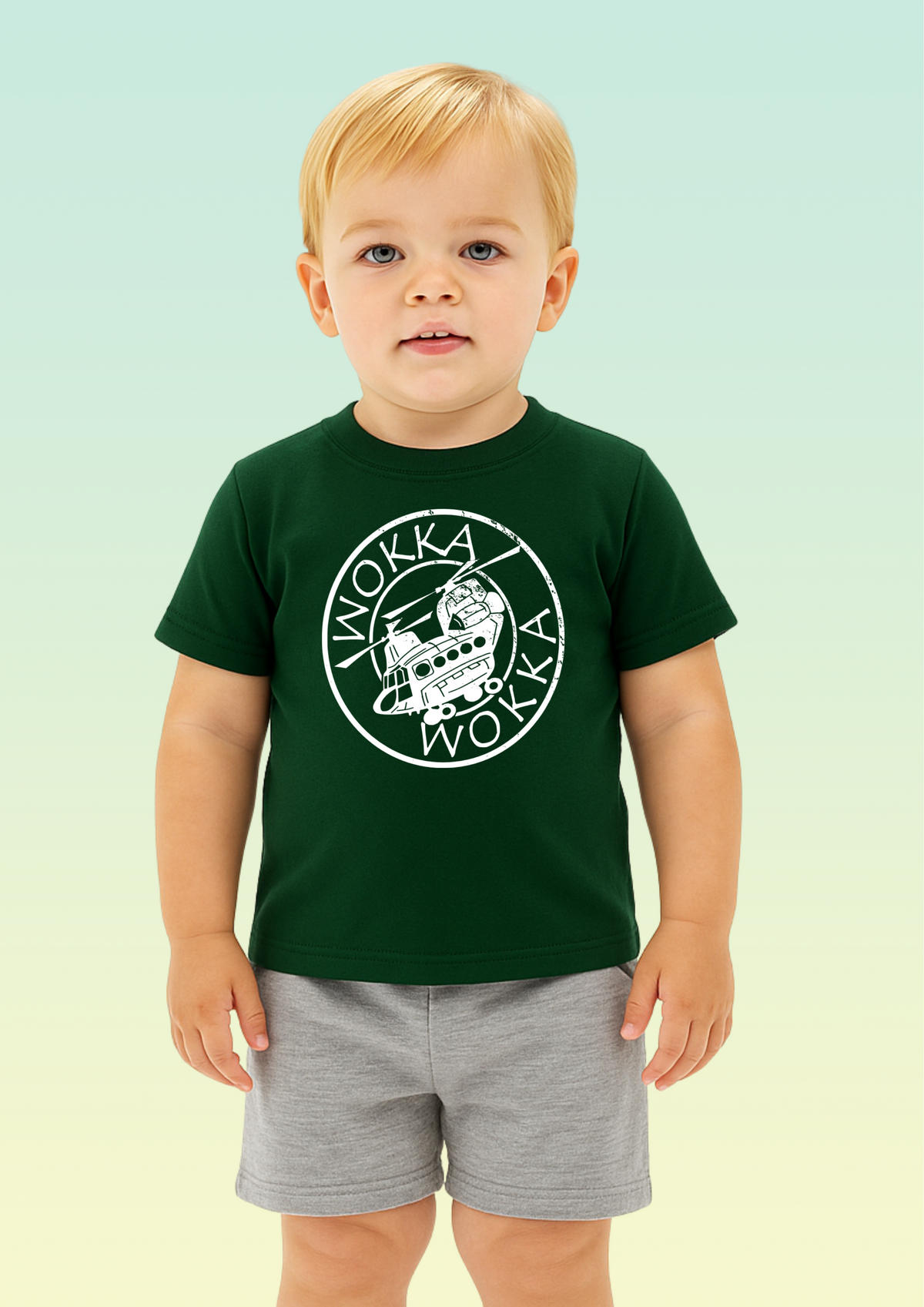 A young toddler with blond hair stands against a pale mint-to-yellow gradient background, wearing a dark green short-sleeve T-shirt with a large white circular 'Wokka Wokka' graphic on the chest and light gray shorts. Exclusive to Flying Teez.