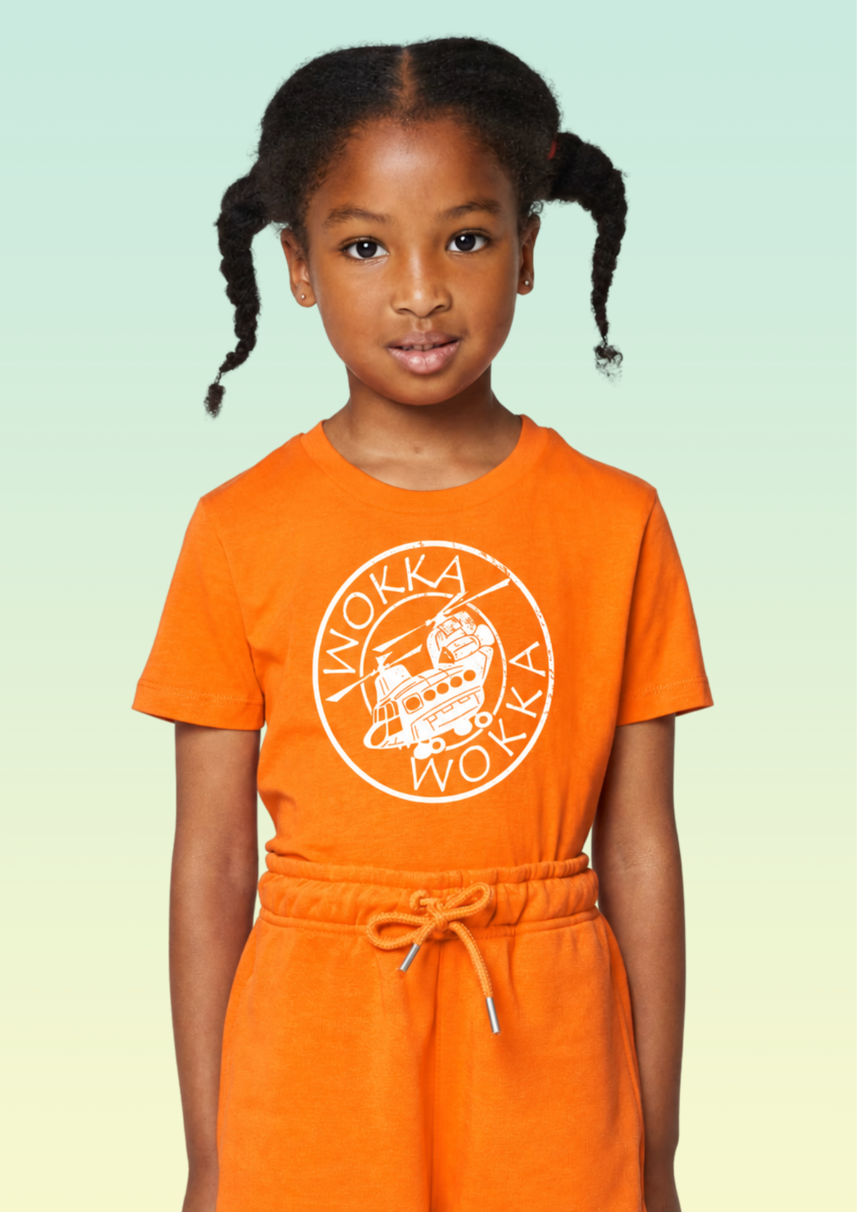 A young girl with braided hair wearing an orange crewneck sweatshirt and matching sweatpants set with a white circular 'WOKKA' graphic on the chest, set against a light green gradient background. Exclusive to Flying Teez.