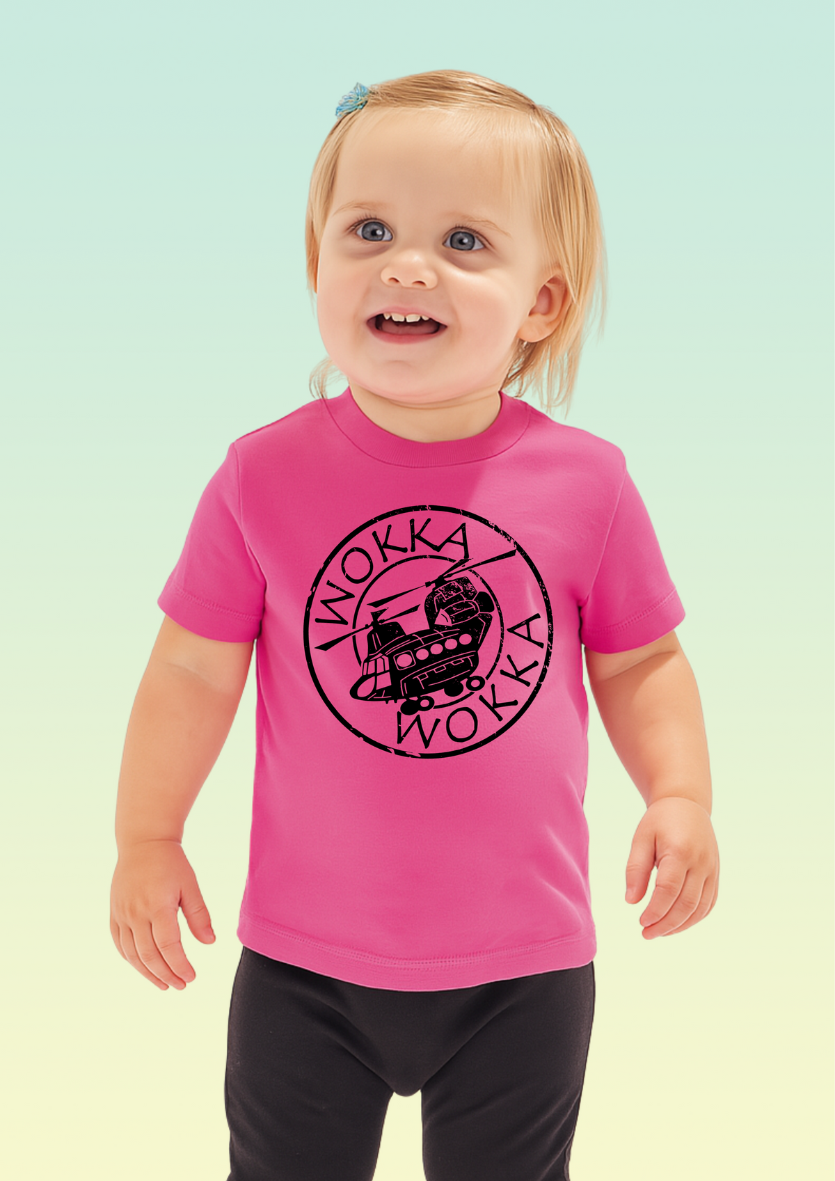 A toddler girl with blonde hair wearing a bright pink t-shirt with a black circular 'Wokka Wokka' graphic on the chest, paired with black pants, set against a light green and yellow gradient background.