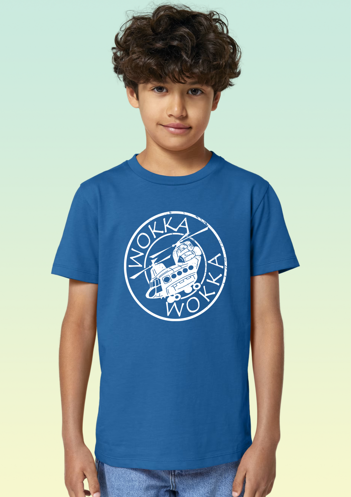 A young boy with curly brown hair wearing a royal blue crewneck t-shirt with a white circular graphic on the chest, set against a light green to pale yellow gradient background. Exclusive to Flying Teez.