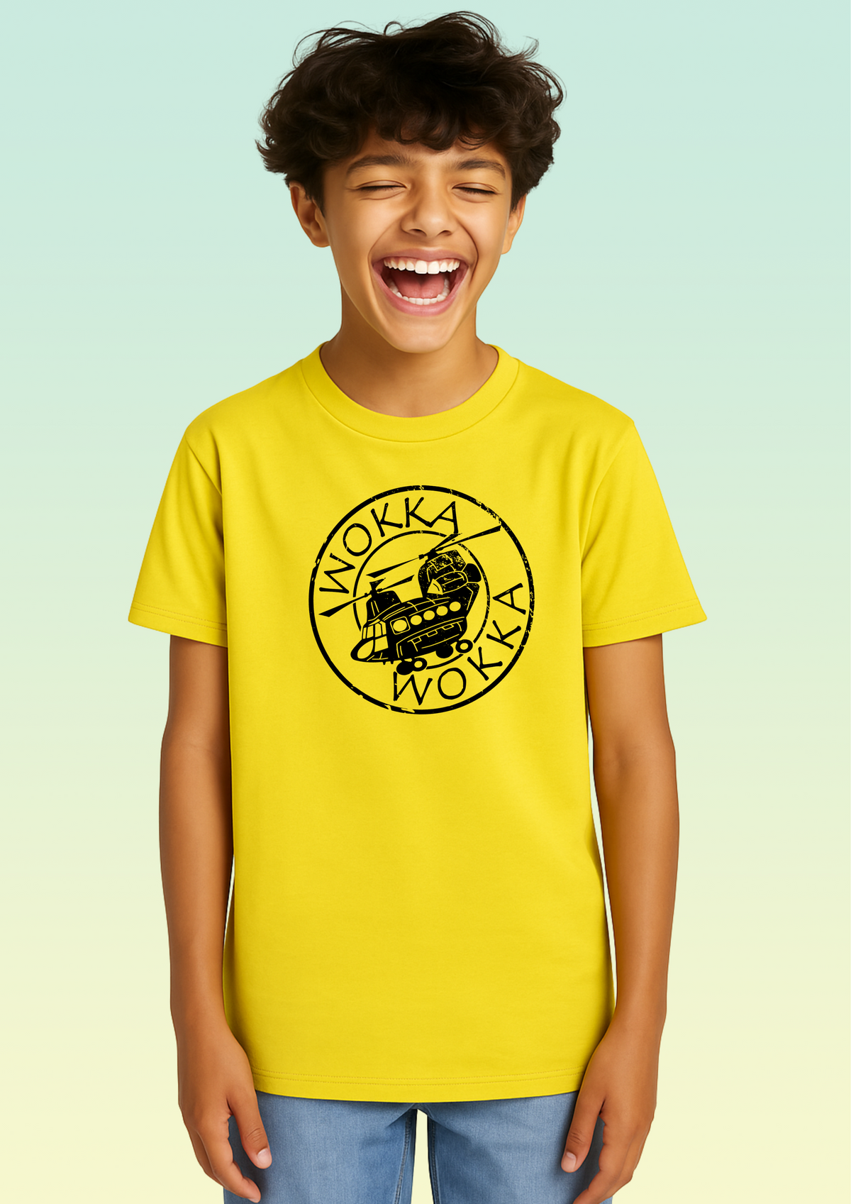 A young boy with dark curly hair is smiling and laughing, wearing a bright yellow crewneck t-shirt with a black circular 'Wokka Wokka' helicopter graphic on the chest, paired with blue jeans. The background is a soft, light green gradient.