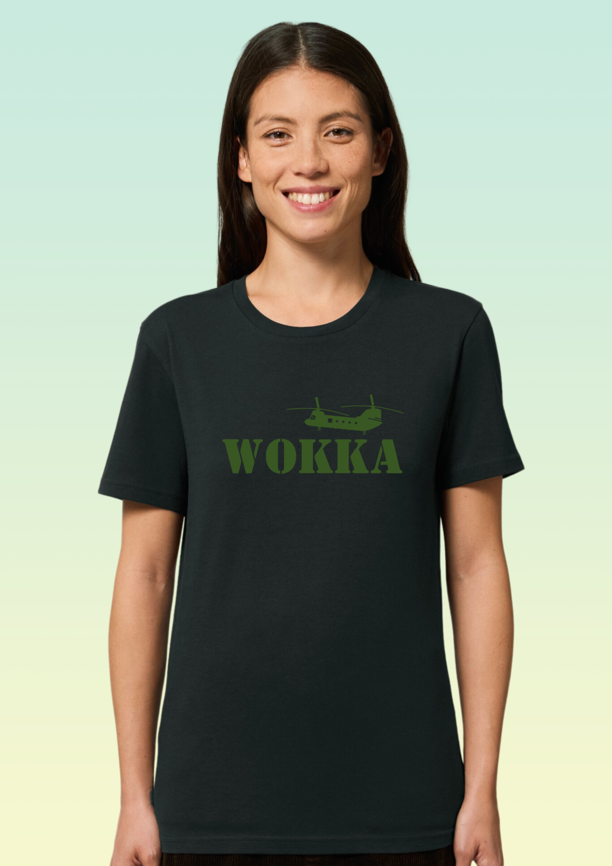 A woman wearing a black short-sleeve crewneck t-shirt with a green graphic of a helicopter and the word 'WOKKA' printed on the chest, set against a light green gradient background. Exclusive to Flying Teez.