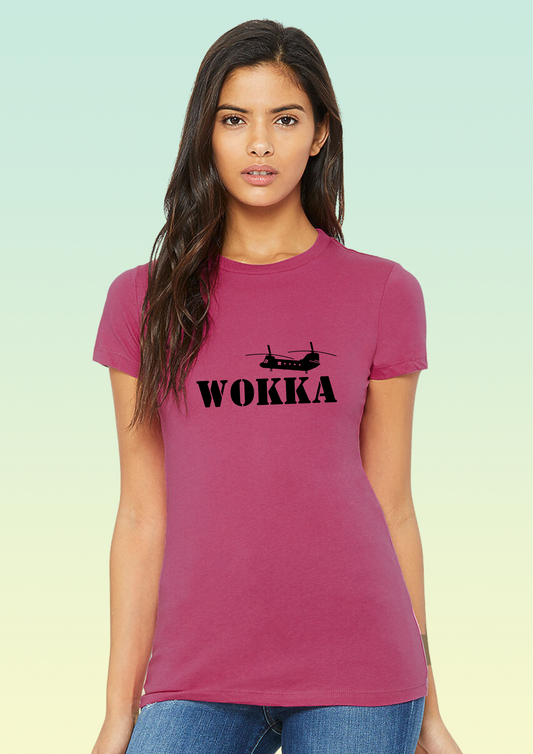A woman modeling a short-sleeve crewneck t-shirt in a dusty rose pink color, featuring a black graphic of a military helicopter and the word 'WOKKA' printed across the chest. She is wearing blue jeans against a light green-to-yellow gradient studio background. Exclusive to Flying Teez.