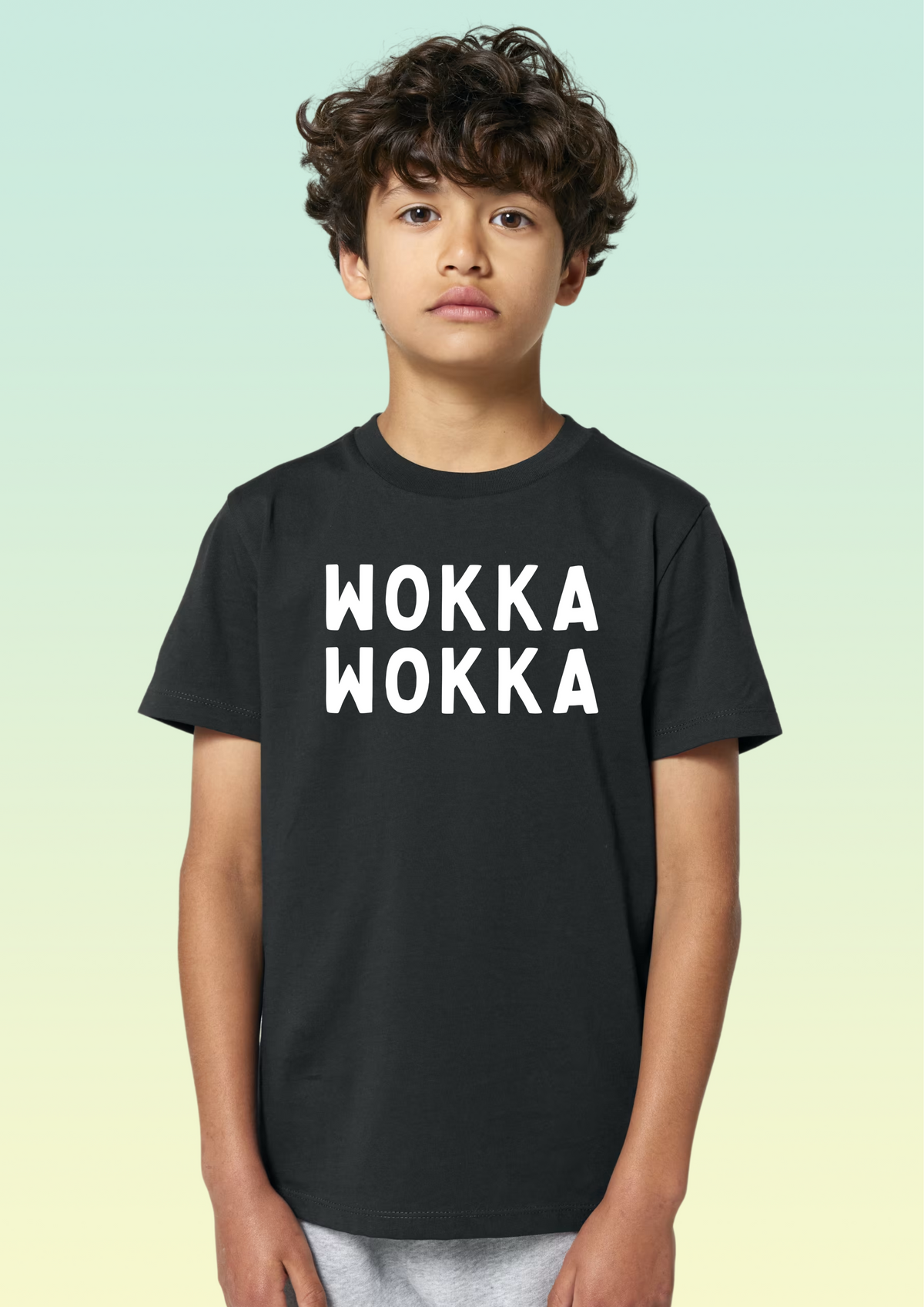 A young boy with curly brown hair wearing a black crewneck t-shirt with 'WOKKA WOKKA' printed in bold white block letters across the chest, set against a light green and yellow gradient background. Exclusive to Flying Teez