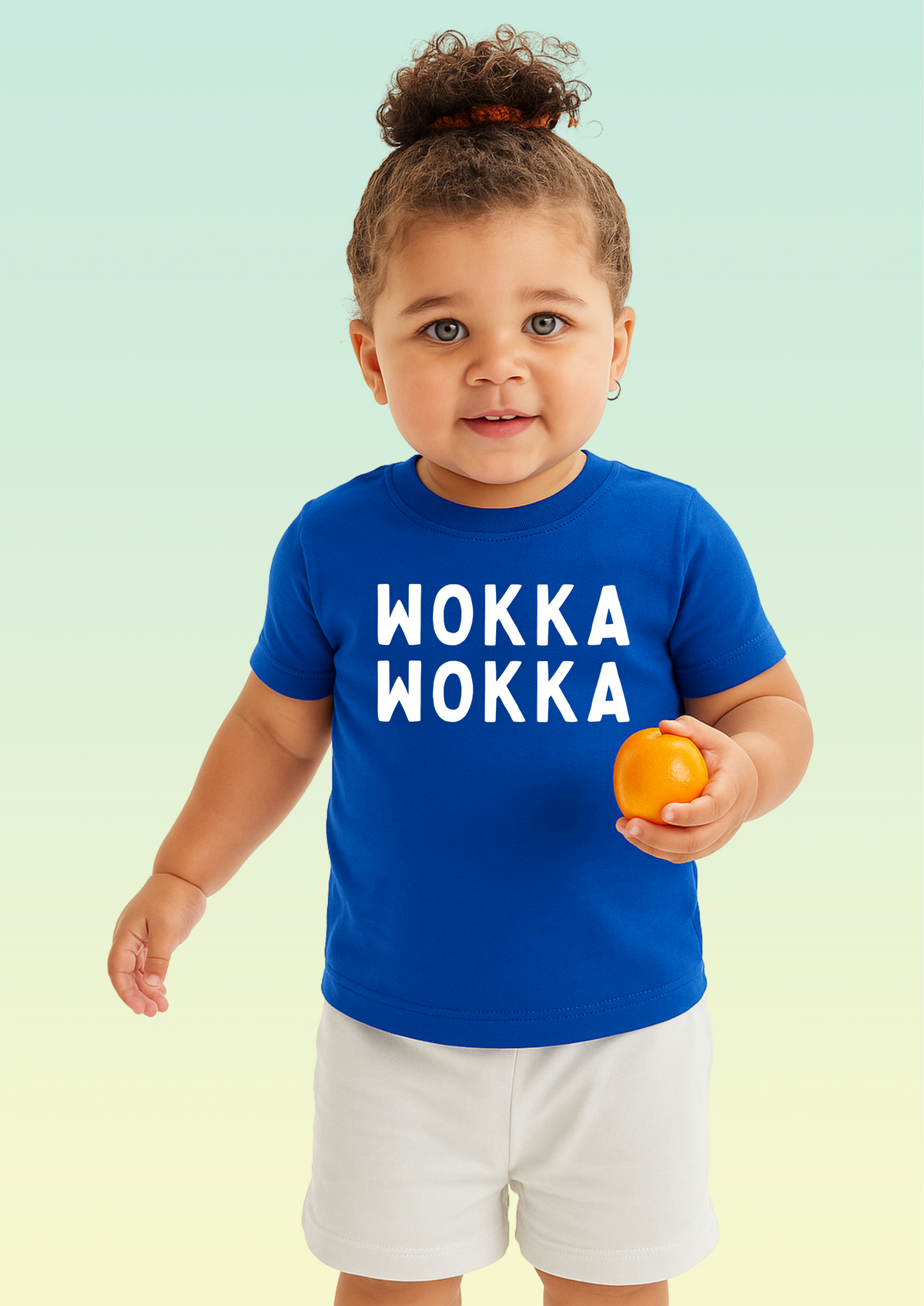 A toddler standing against a pale mint-to-yellow gradient background, wearing a bright blue t-shirt with large white text reading 'WOKKA WOKKA' and white shorts, while holding a small orange. Exclusive to Flying Teez.