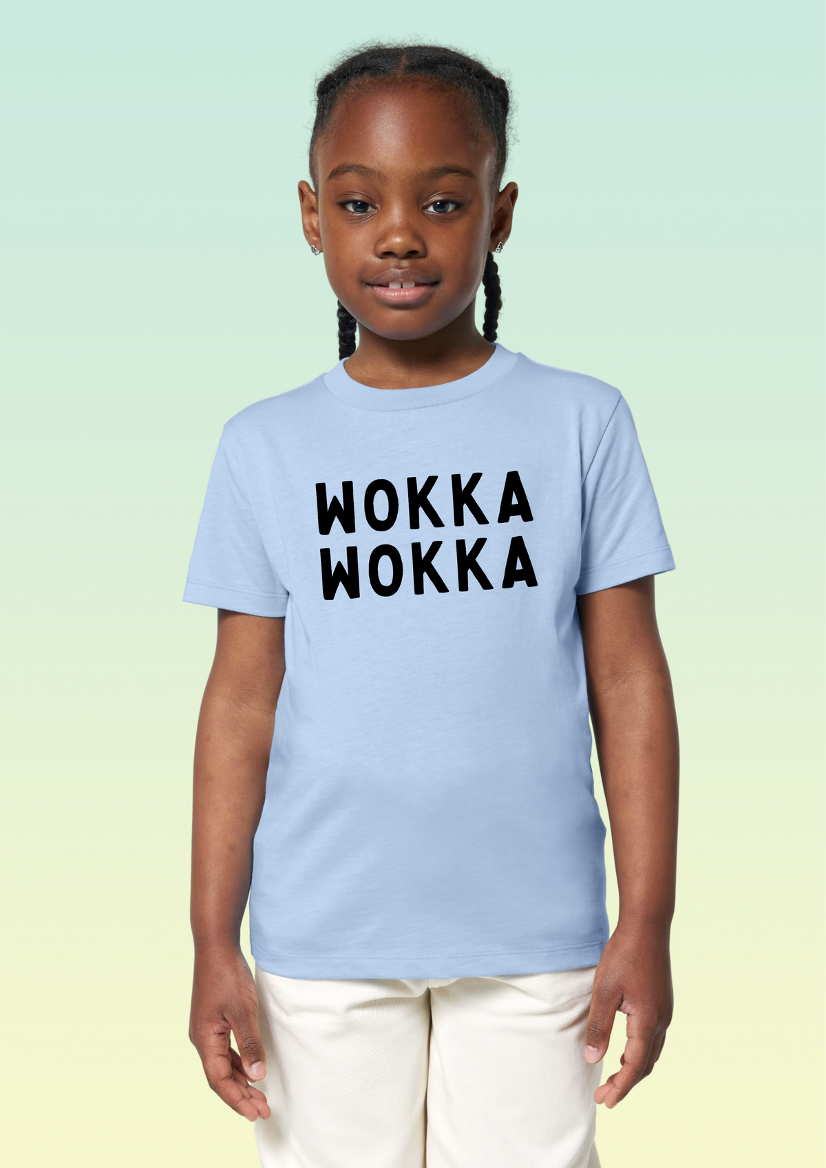 A young girl with braided hair wearing a light blue t-shirt with the words 'WOKKA WOKKA' printed in black across the chest, paired with cream pants, set against a light green gradient background. Exclusive to Flying Teez.